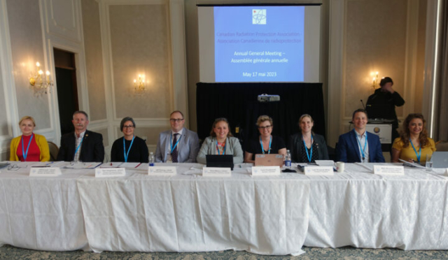 2022–2023 Board of Directors at the AGM. a row of people sit behind a long table with a white table cloth. On a screen behind them is a the SCPR logo and the words Annual General Meeting..