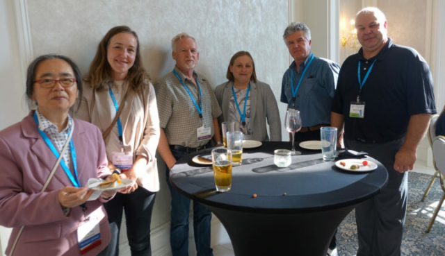 From left to right: Jing Chen, Lauren Bergman, Cody Cuthill, Diana Moscu, Rick Kosierb, and Sylvain Faille. Six people standing in a row behind a reception table with food and drinks.