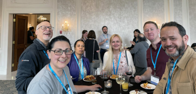 From left to right: Ralph Bose, Tanya Vlaskalin, Vani Ranganathan Donata Chaulk, Justin Symons, Fouad Bacher. A group of people sit around a table with food and wine. They are looking at the camera. Another table of people can be seen in the background.