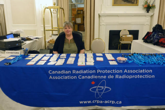 Sue Singer (secretariat) at the registration desk a woman sits behind a table with a blue table cloth. The CRPA logo is printed on the front of the tablecloth. On top of the table are name badges and laniards