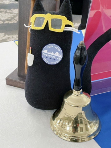 Schrodinger repping the next CRPA conference to be held in Edmonton in 2024! We hope to see you all there. A stuffed cat with a button promoting the Edmonton conference sits on a table next to a gold bell.
