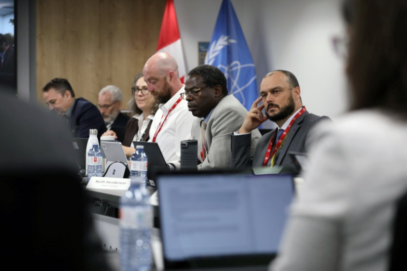 A group of people sitting at a table in front of a Canadian flag and an International Atomic Energy Agency flag. The is one woman and five men in the picture.