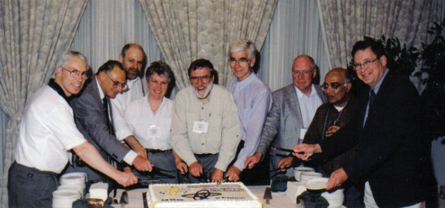 CRPA presidents cut into the 20th Anniversary cake at the conference in Saskatoon, SK, in 1999. Left to right, Mike Haynes (98–99 president), Denis Brown (98–99 president elect), Gary Kramer (97–98 president), Catriona Maloney (95–96 president), Wayne Greene (83–84 president), Richard Osborne (79–80 president), John Johnson (84–85 president), Robi Chatterjee (90–91 president), Harry Johnson (99–00 president elect). A row of 9 people (mostly men), cutting a large cake that is in the middle of the table in front of them