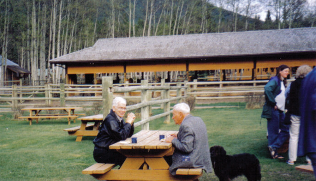 Richard Osborne and Tony MacKay (archivist) chatting at Boundary Park Ranch in Kananaskis, AB, at the CRPA conference in 2003. 2 older men sitting at a picnic table outside in a mountainous area. A small group of people are standing at the right side of the picture.