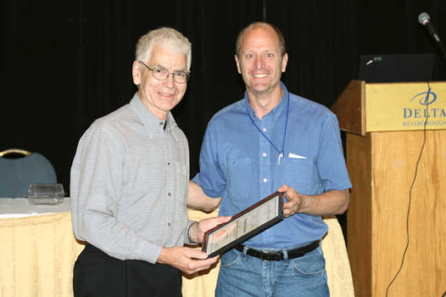 Richard Osborne receiving the first Honorary Lifetime Membership plaque from President Gary Wilson (2008). A man with grey hair and glasses wearing a grey dress shirt and black pants receiving a plaque from a man wearing a shirt-sleeved blue button-up shirt and jeans