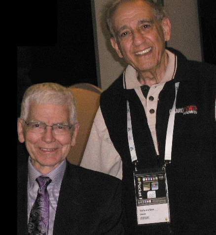 Richard Osborne with Nick Sion, who was another of CRPA’s earliest members a man with grey hair and glasses in a suit and tie sitting in a chair while another grey-haired man wearing a polo shirt with a conference laniard stands beside and slightly behind him