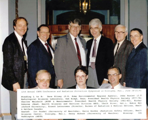 a row of 6 older white men wearing suits and ties standing behind 3 people who are seated in front of them: an older blind man on the left, a dark haired woman in the middle, and a dark-haired man on the right. At the bottom of the picture is a white box with typed caption