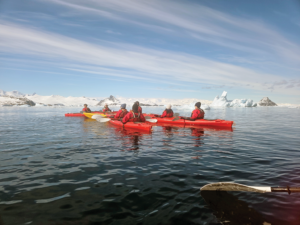 People in red kyaks on dark water. THe shore is snow-covered. The sky is bright and steaked with clouds.