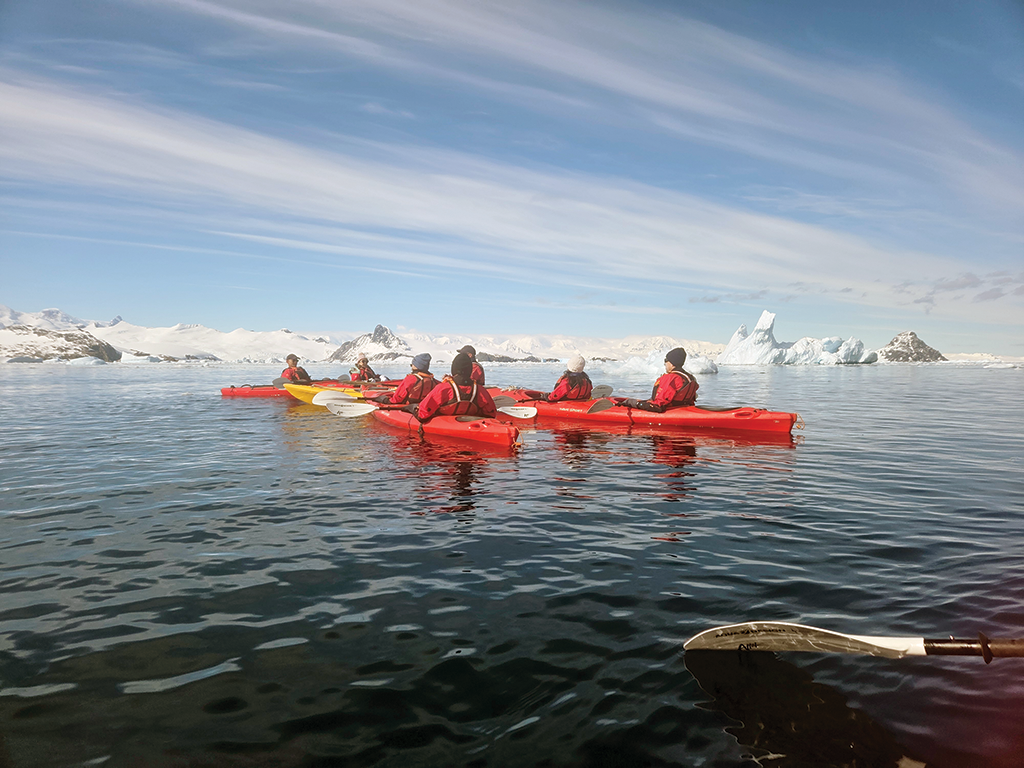 People in red kyaks on dark water. THe shore is snow-covered. The sky is bright and steaked with clouds.