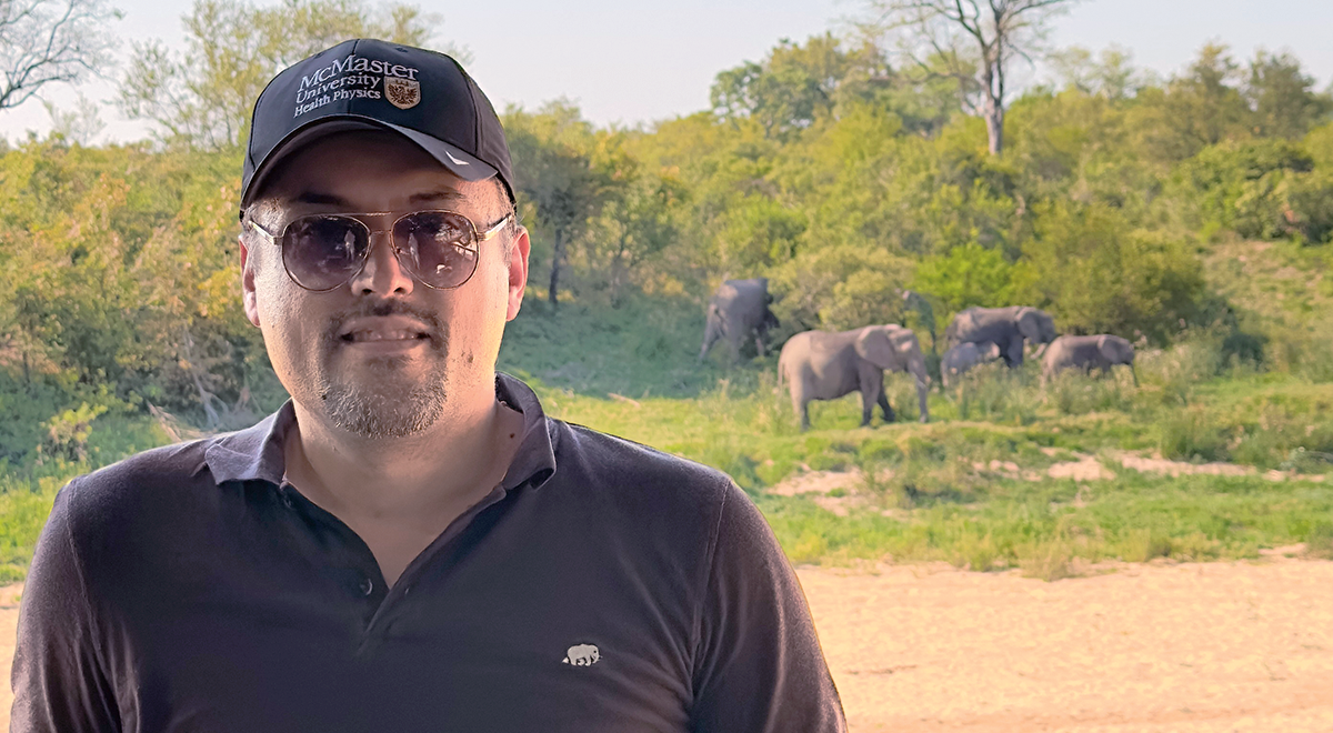 A man standing on the front left side of the image. There are trees behind him and a small group of elephants between him and thre trees on the right side.